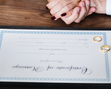 Hands clasped above a wedding certificate on a wooden table, with two rings beside it