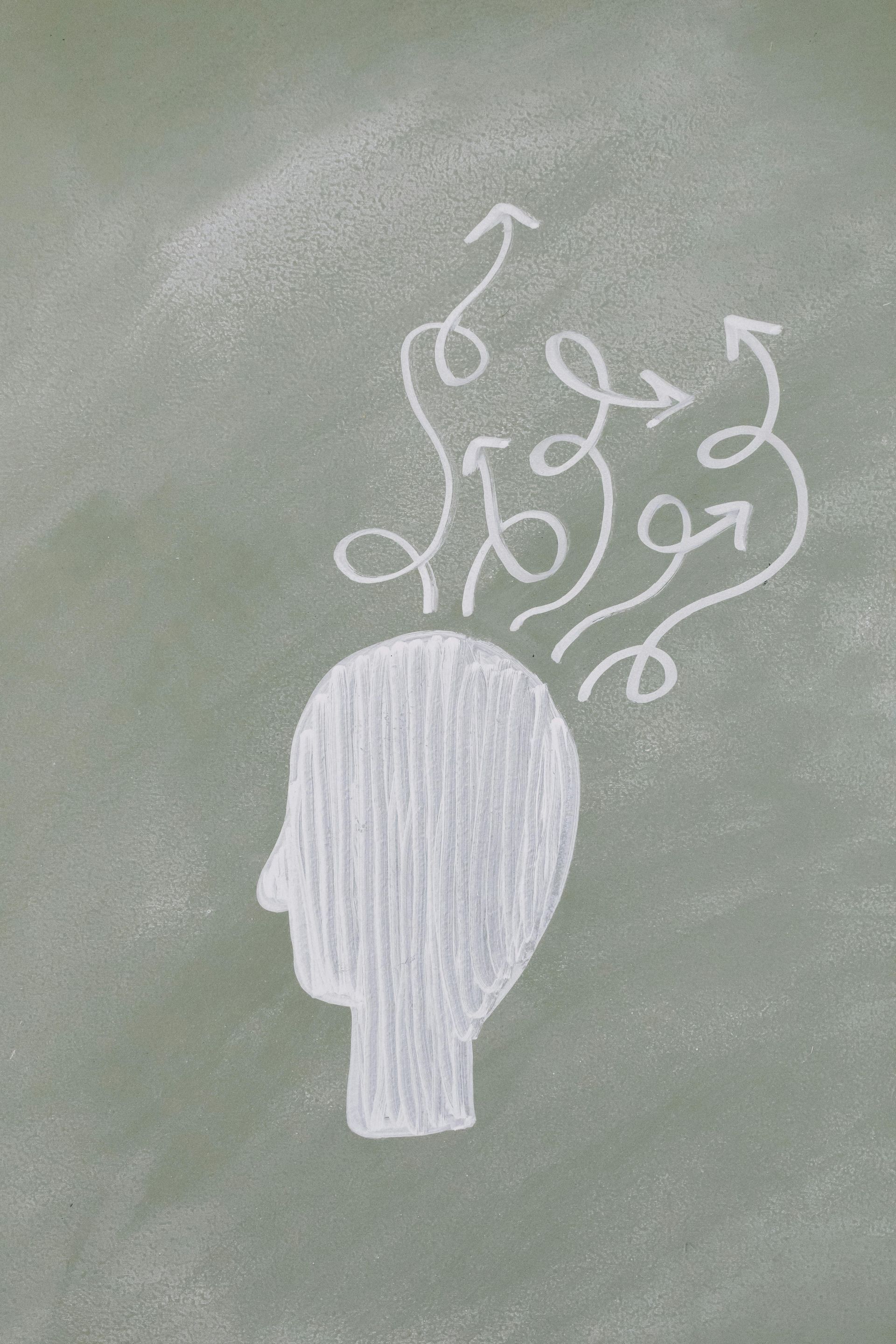 White chalk drawing of a head with arrows and swirls emanating from it on a green chalkboard, representing thought or ideas.