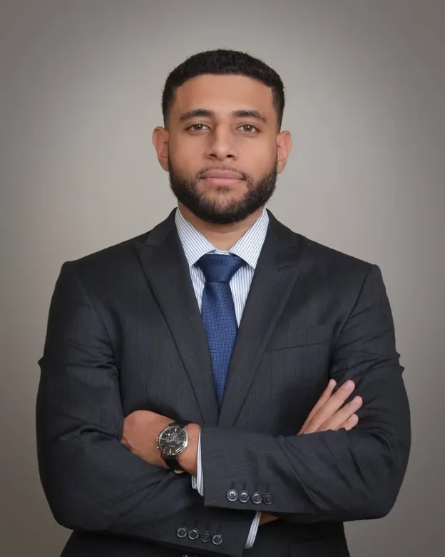 Man in dark suit and blue tie standing with arms crossed against a gray backdrop