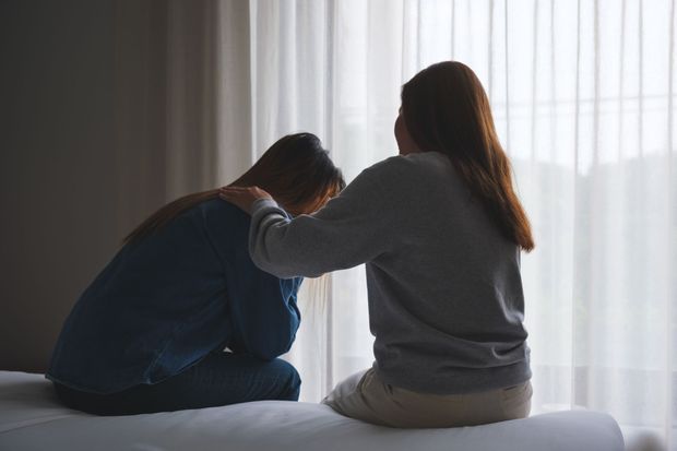 Two people sitting on a bed, one comforting the other by placing a hand on their shoulder.