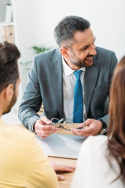 Man in gray suit speaking with colleagues at a meeting table, smiling and holding glasses