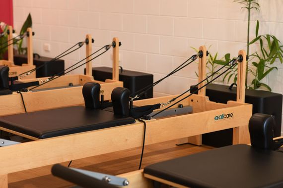 Pilates reformers in a studio, light wood with black cushions and straps, near a brick wall and plants.