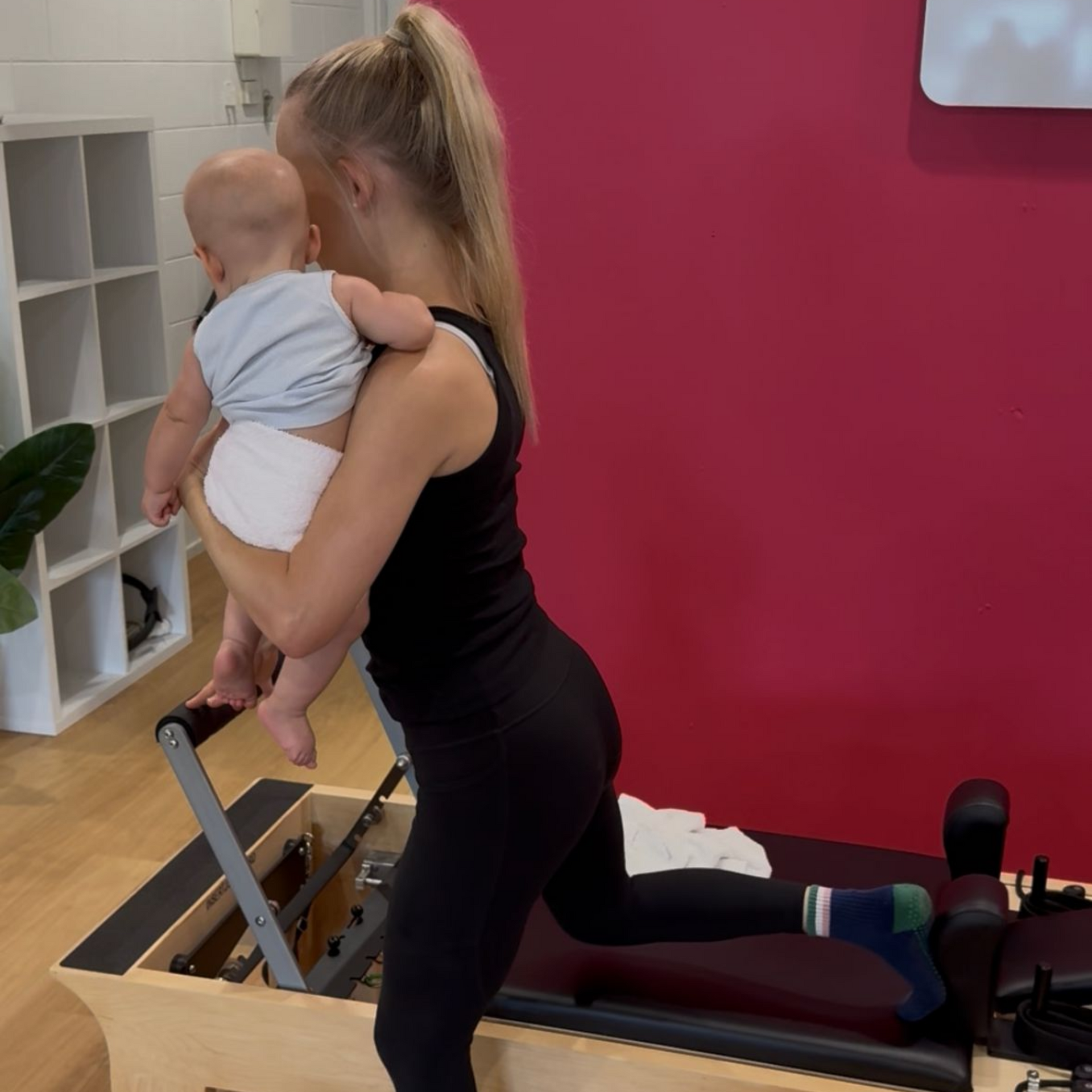 Woman exercising on a Pilates reformer while holding a baby in a bright studio.