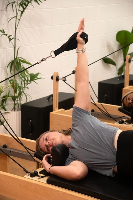 Woman doing Pilates exercise on a reformer machine, arm raised and holding strap, lying down.