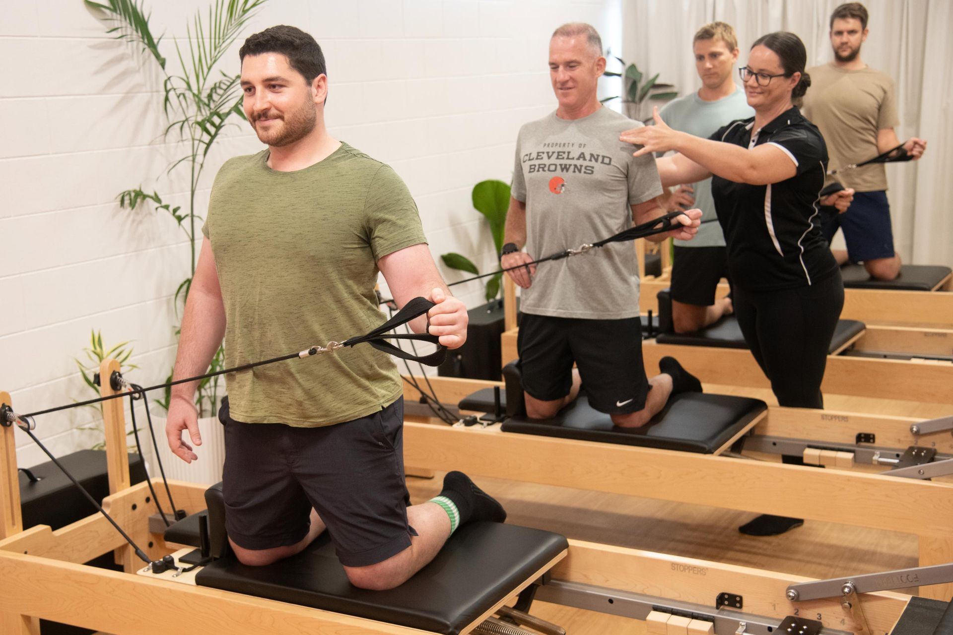 People in a fitness studio exercise on Pilates reformers while a trainer provides guidance.