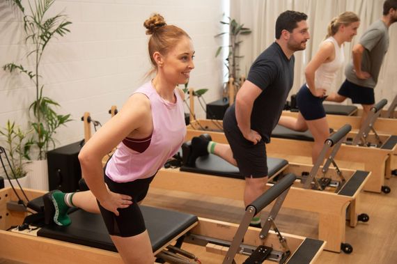People doing Pilates exercises on machines in a bright studio, smiling.