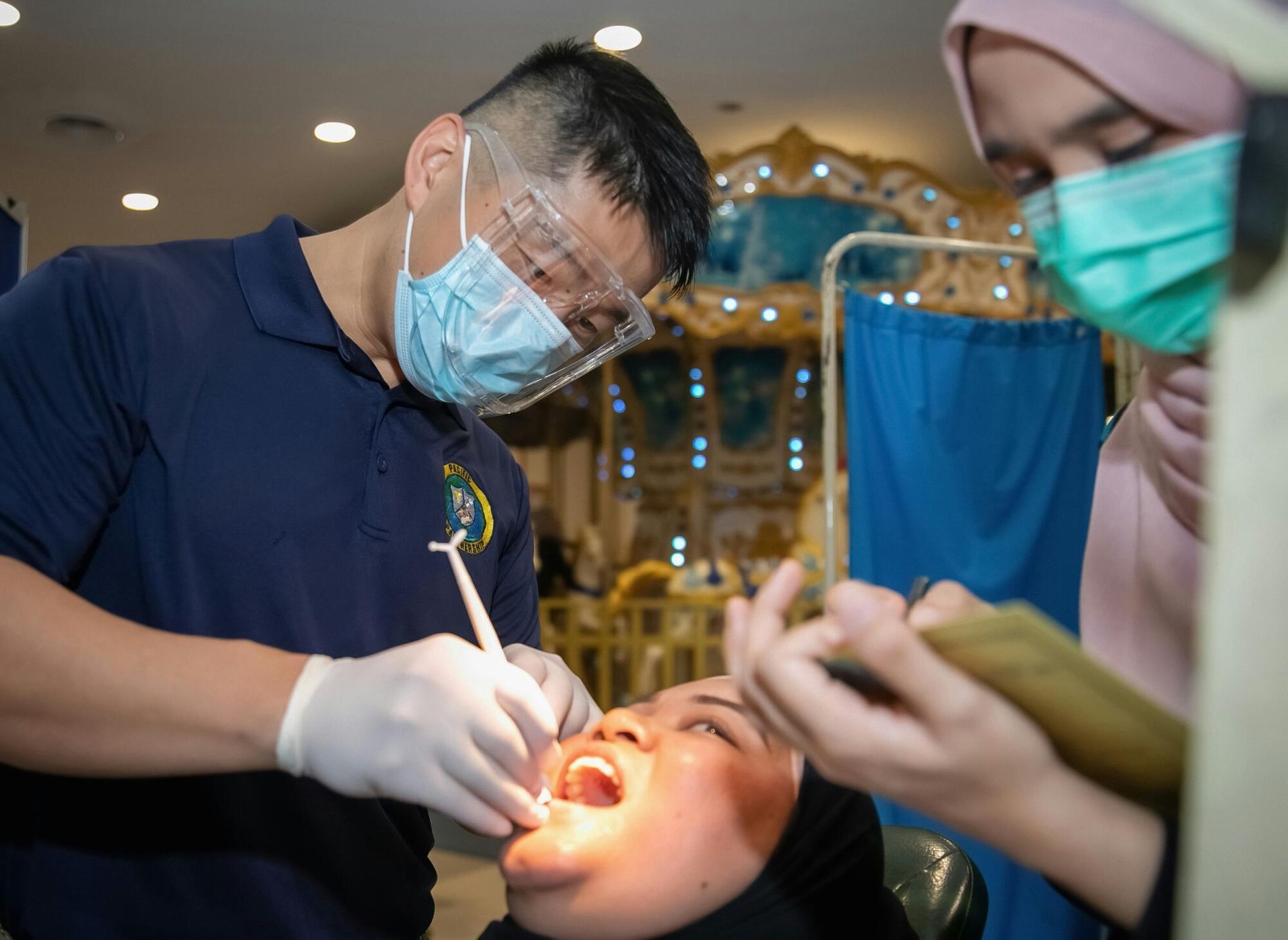 A dentist with a mask and face shield examines a patient's mouth while an assistant takes notes.