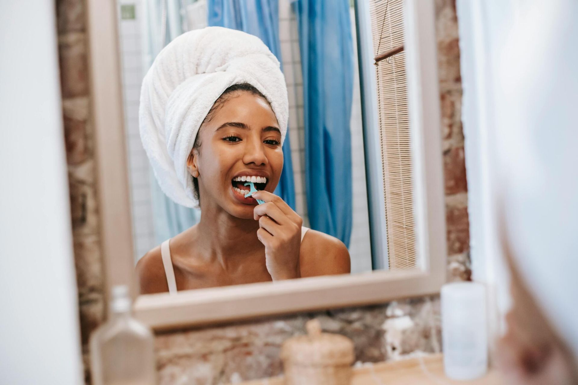 Person brushing teeth in a bathroom mirror, wrapped in a white towel, with blue curtains behind.