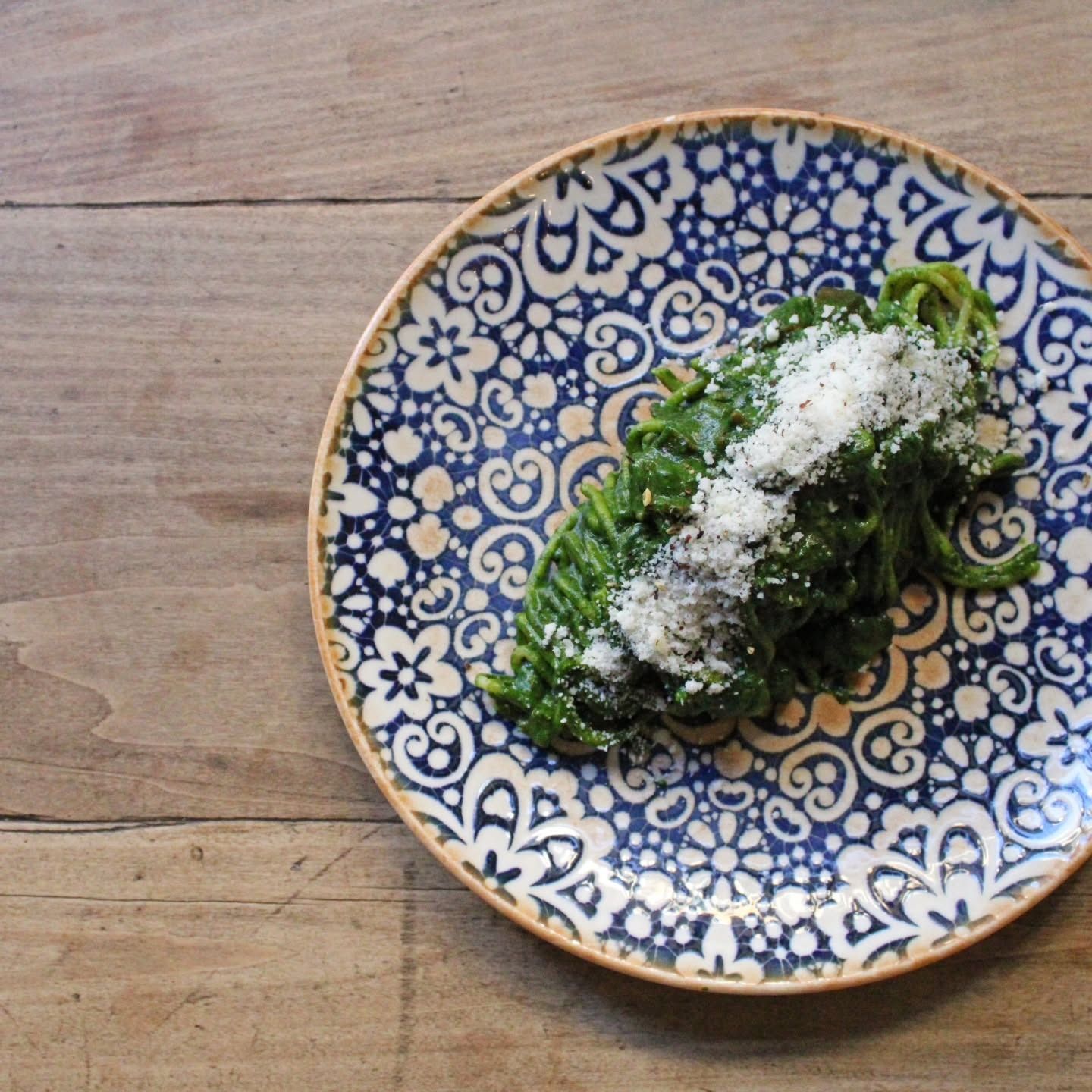 Pasta agli spinaci con formaggio grattugiato su un piatto decorato con motivi blu e bianchi, appoggiato su una superficie di legno.
