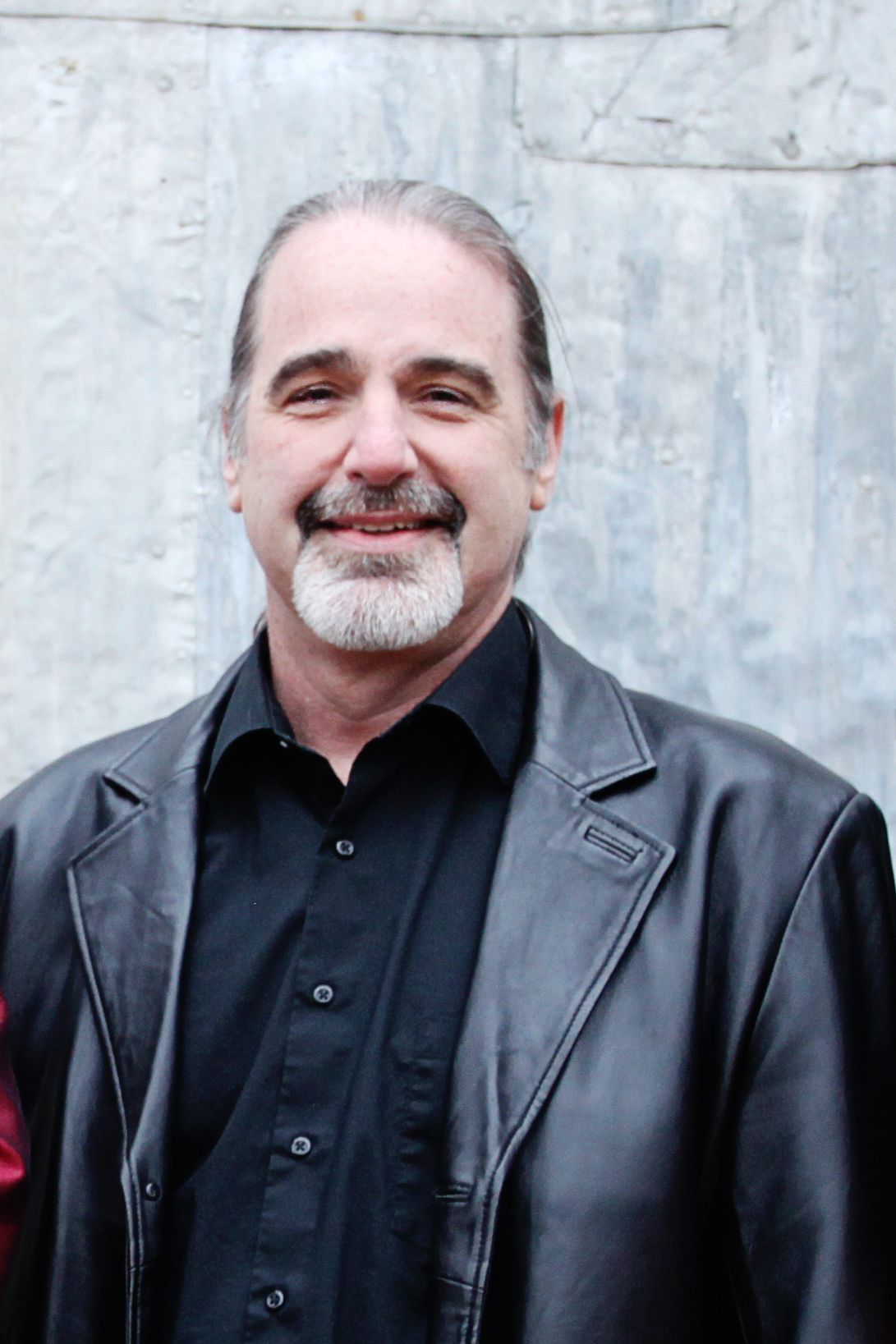 A man with a beard is wearing a black shirt and a black leather jacket.