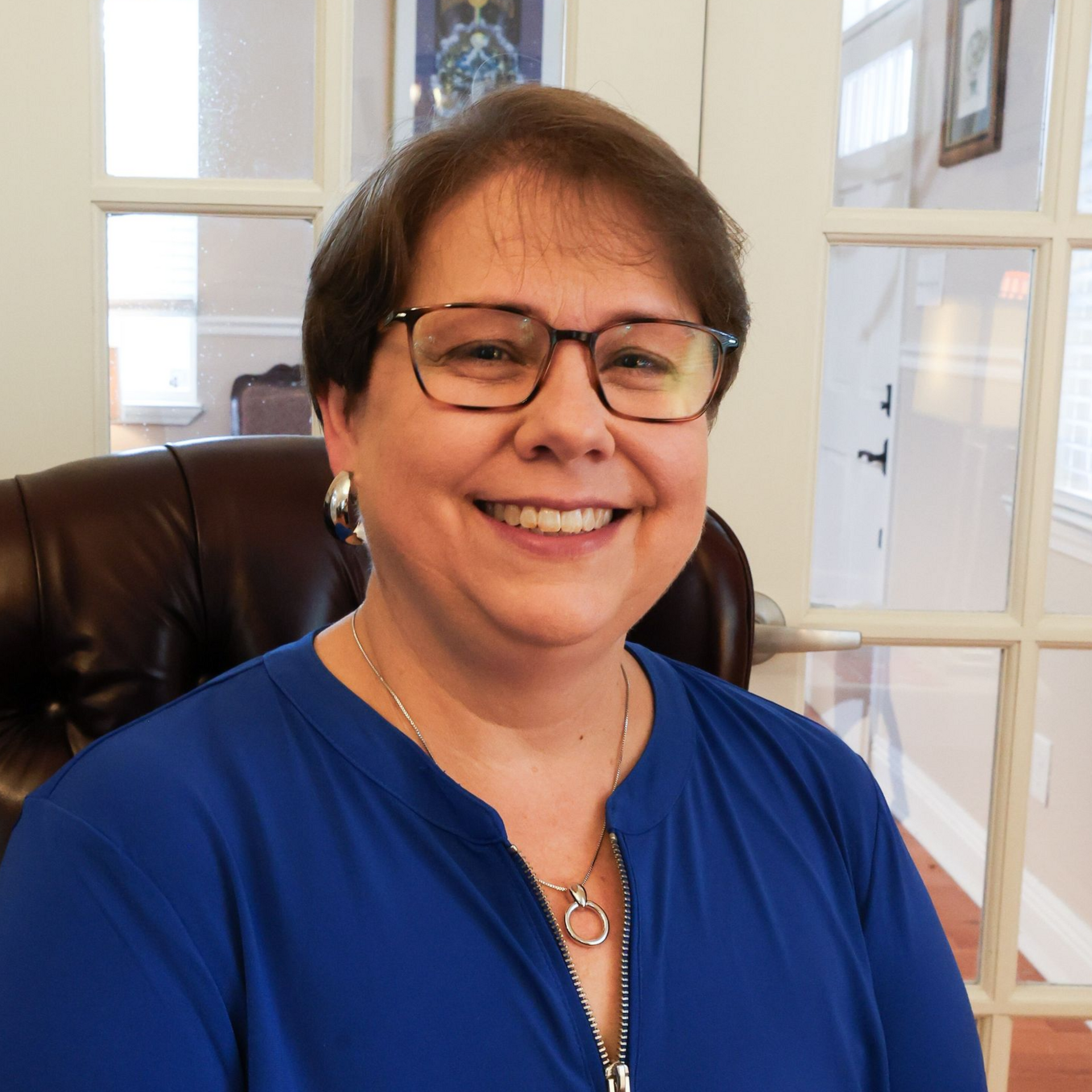 Smiling person in glasses and blue top seated indoors by a window