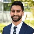 Man in navy suit, white shirt, and patterned tie smiling outdoors.