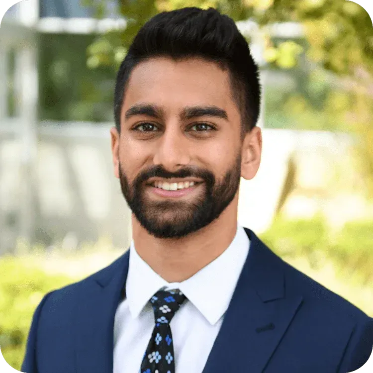 Man in navy suit, white shirt, and patterned tie smiling outdoors.