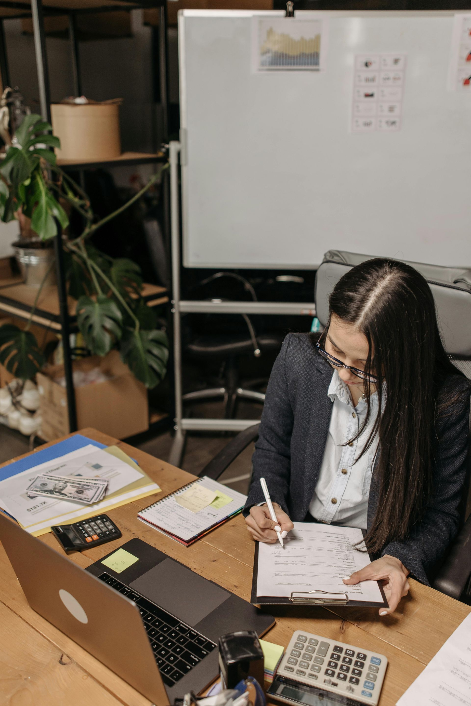 Person writing at a desk in an office with a laptop, papers, and a whiteboard in the background