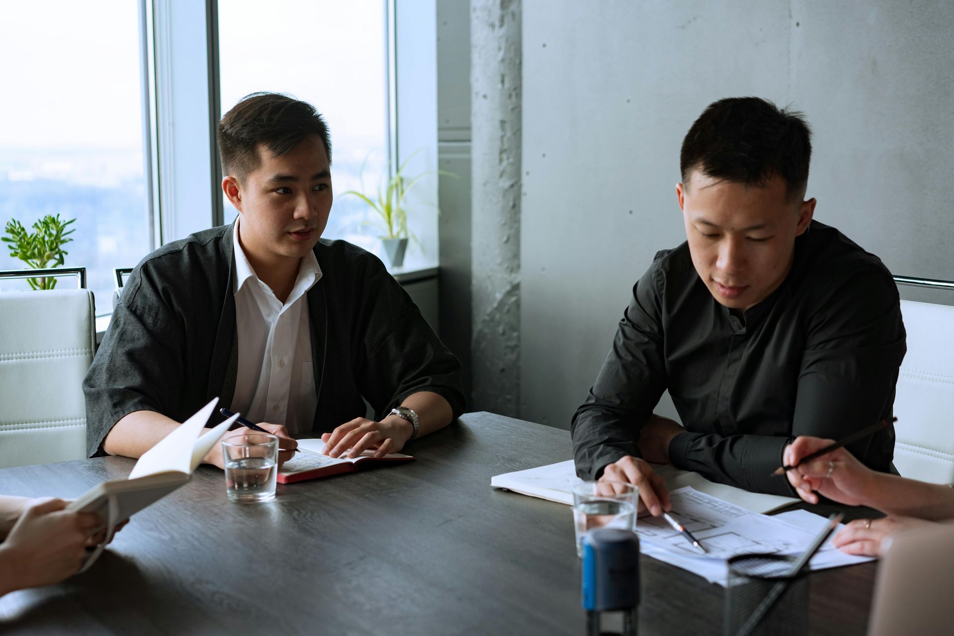 Two men in dark shirts review papers at a table in a bright office meeting room.