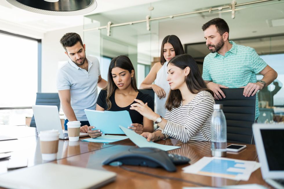 Man points at laptop screen, discussing with woman on a couch in a modern office.
