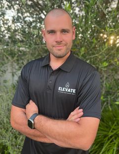 Man in black Elevate polo stands with arms crossed outdoors in front of leafy greenery