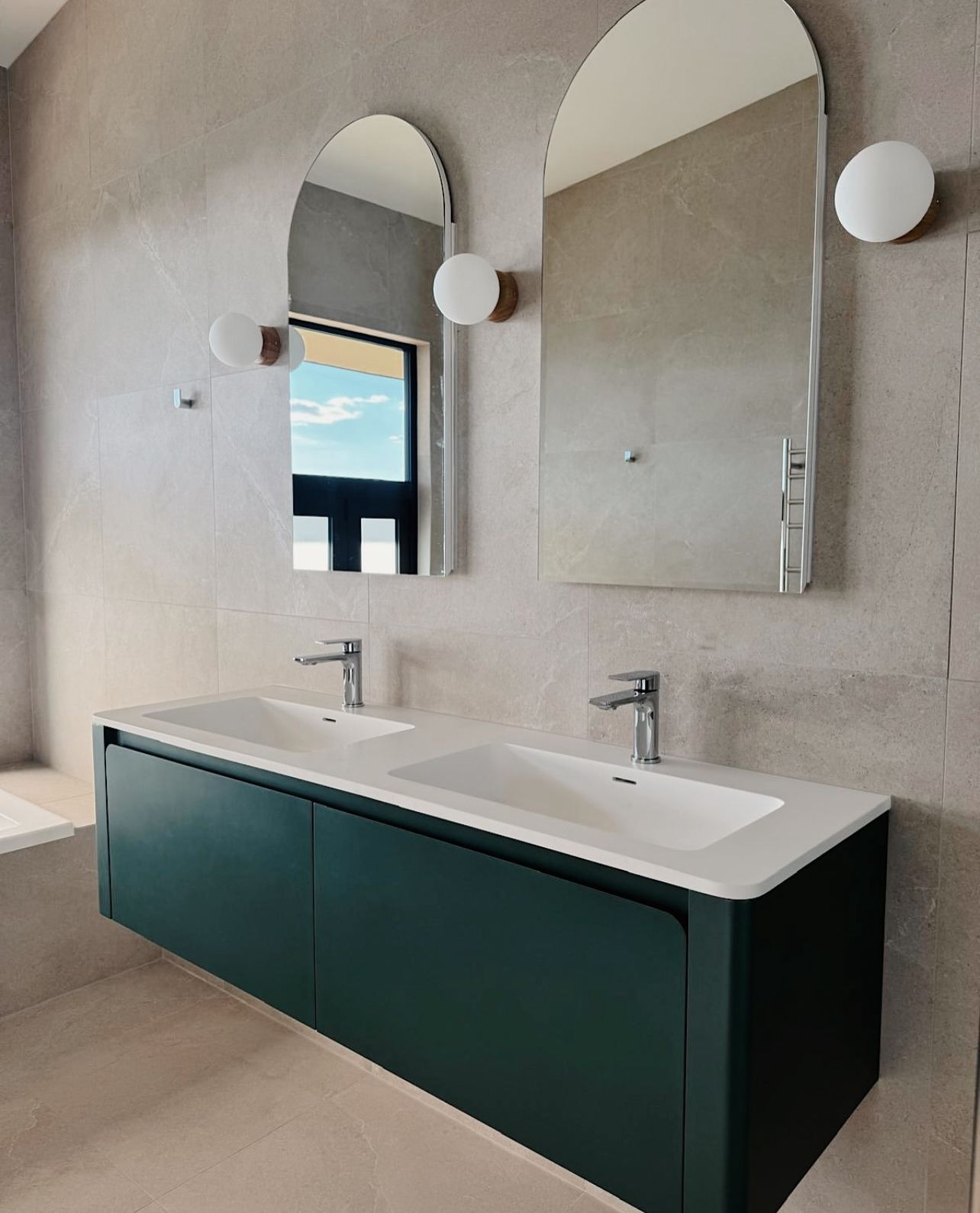 Modern bathroom vanity with dark green cabinets, white sink, and arched mirrors on a textured wall