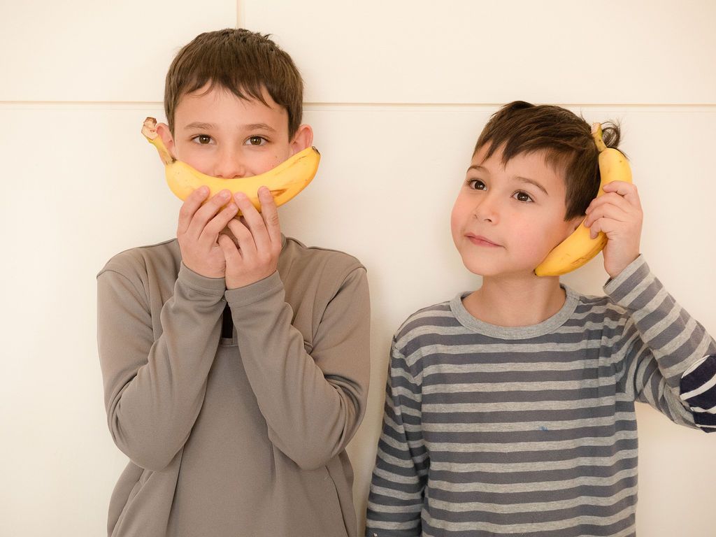 Two boys standing against a wall and being goofy holding bananas.