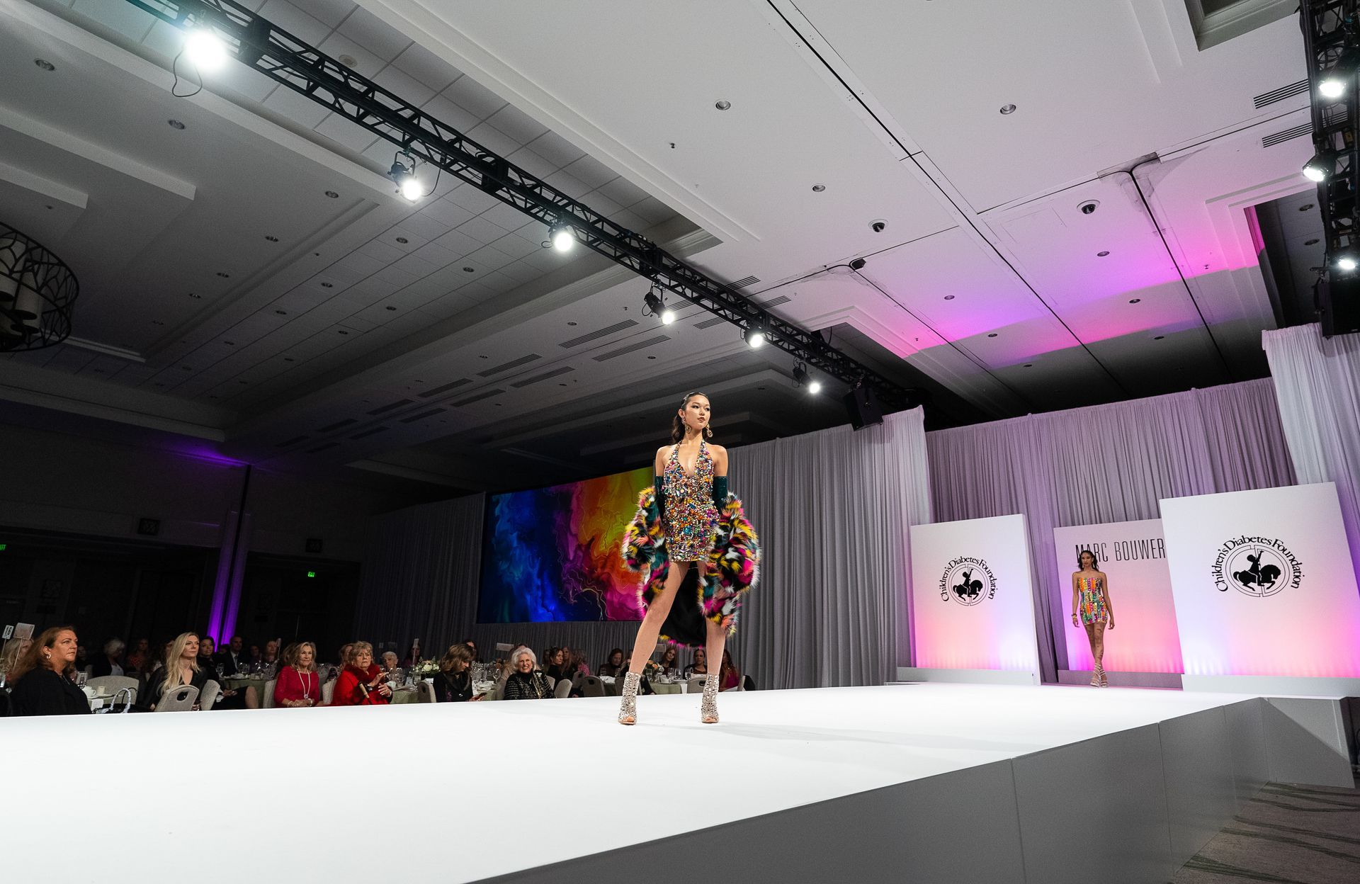Model on runway in multi-colored beaded cocktail dress with multi-colored faux fur coat.