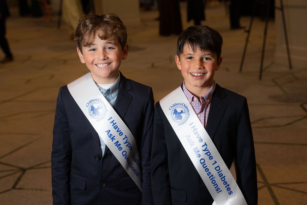 two young white boys who are T1D advocates, wearing a sash asking people to ask them questions about t1d