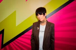 A woman in a suit is standing in front of a pink background.