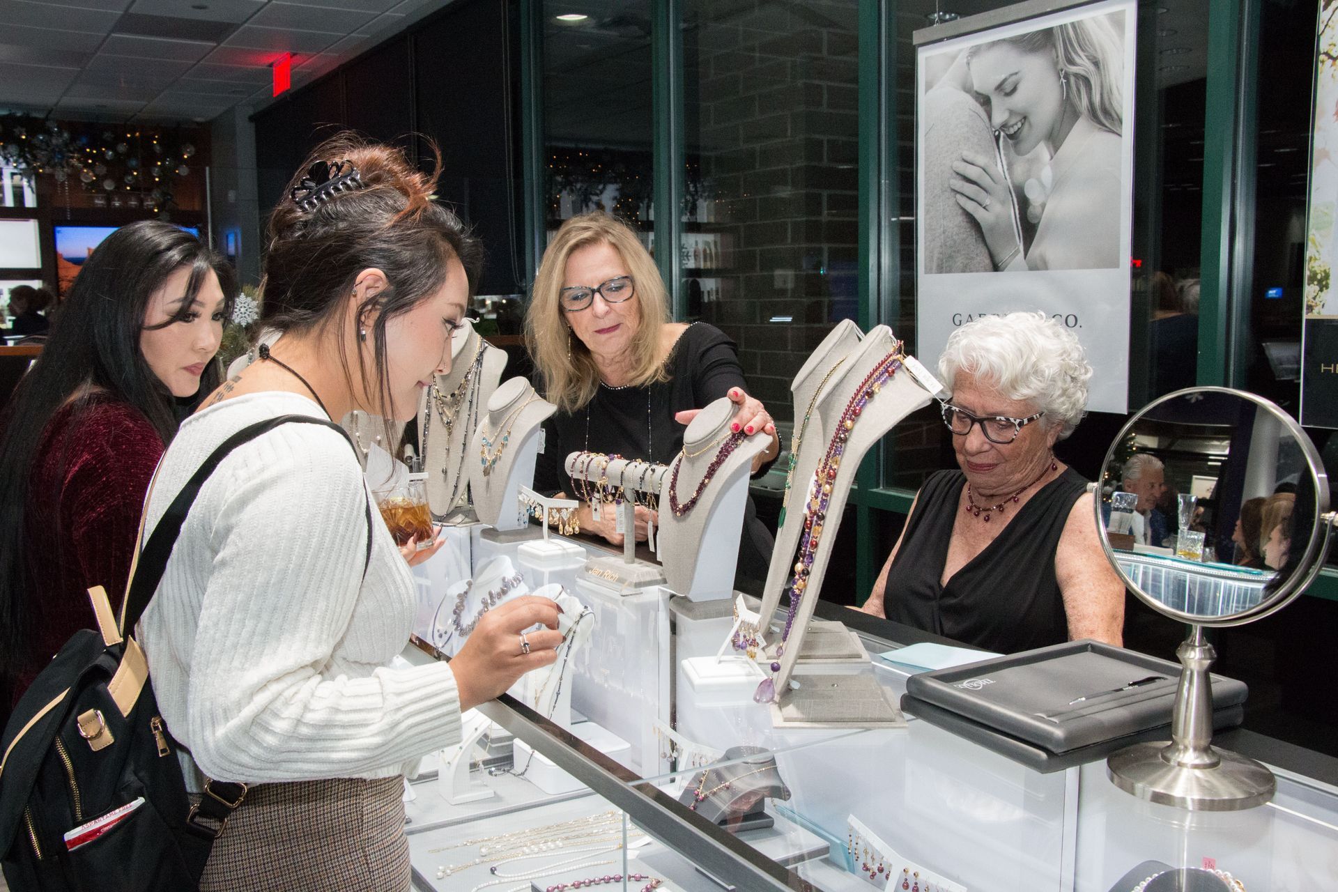 Three women browsing jewelry displayed on a counter inside a store. 
