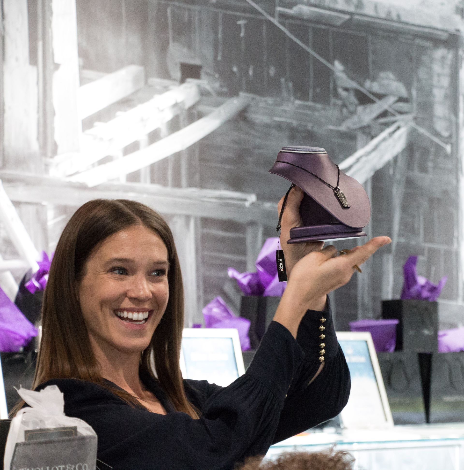 A woman smiling and holding up a purple necklace stand inside a store or showroom. 