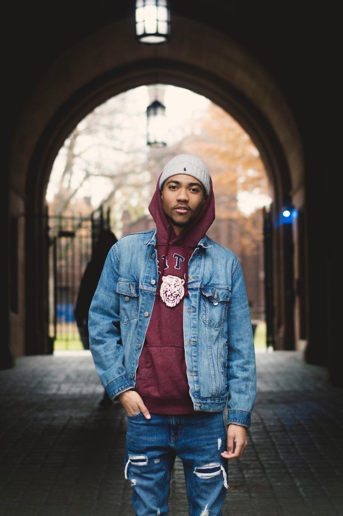 A man wearing a denim jacket and a hoodie is standing in an archway.