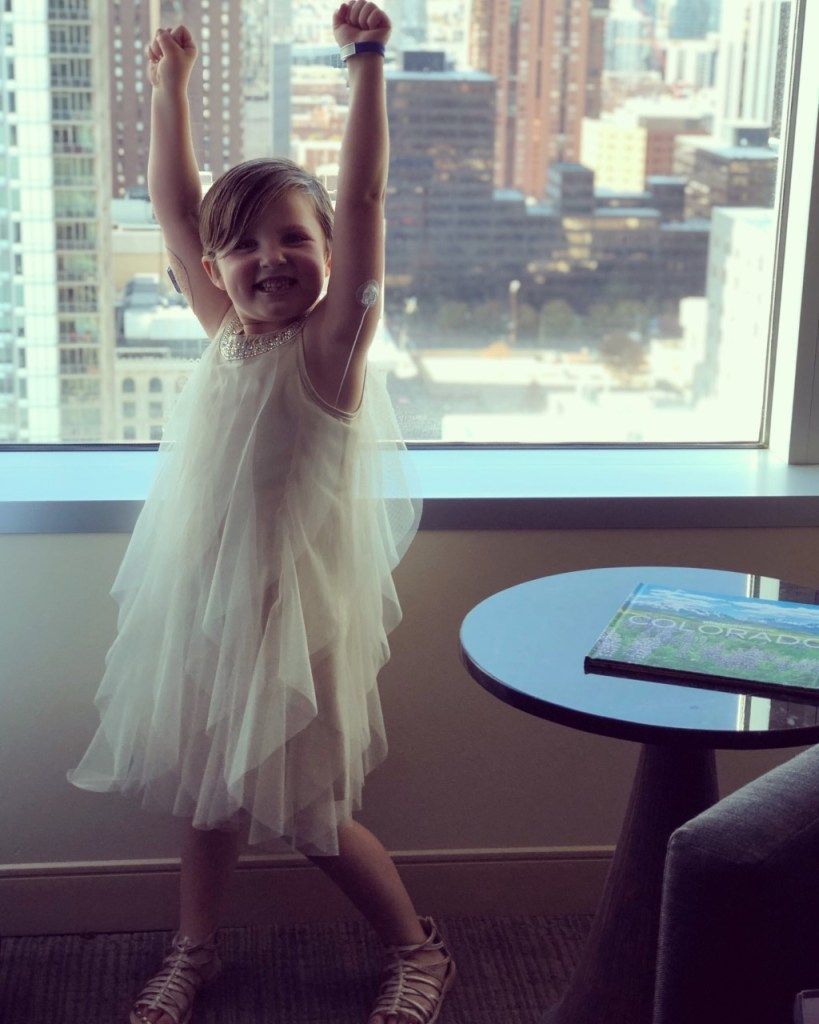 Frankie in a white dress is standing in front of a window with her arms in the air and a CGM on her arm