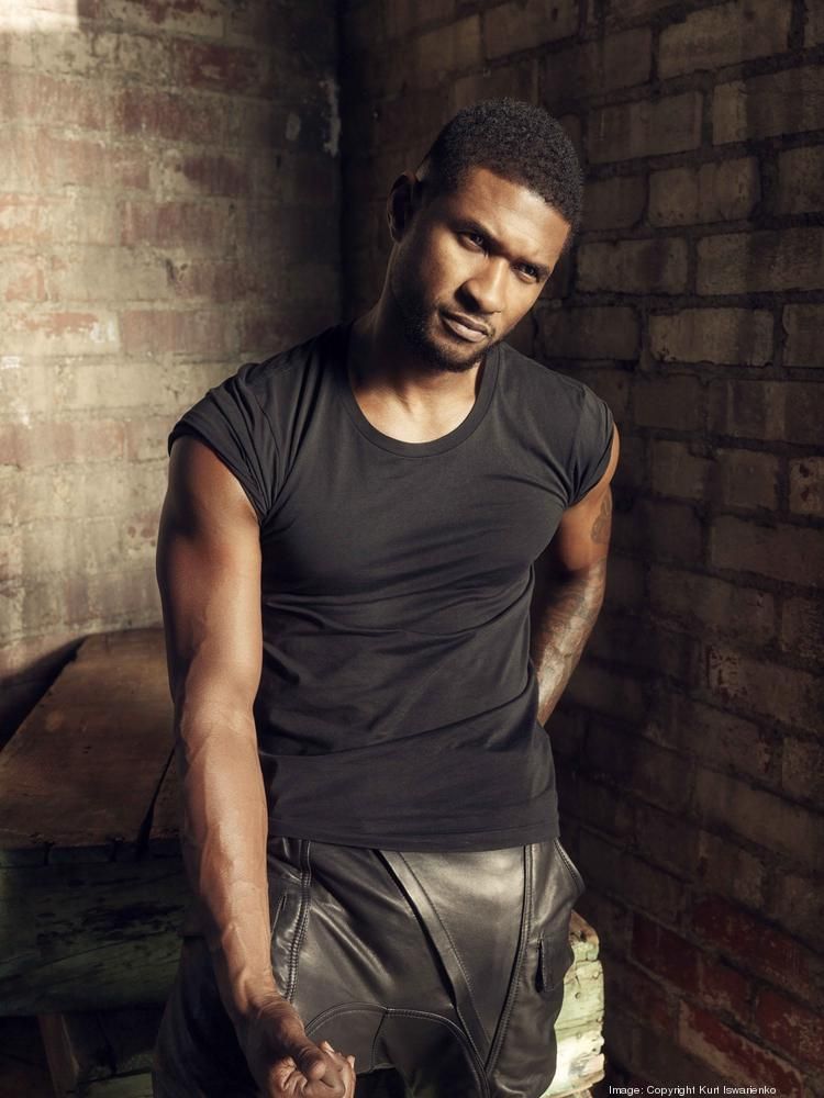 A man in a black shirt and leather shorts is standing in front of a brick wall
