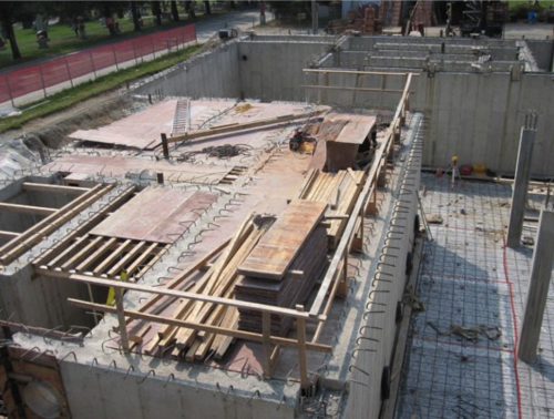 Chantier de construction, fondations en béton, planches de bois et barres d'armature visibles.