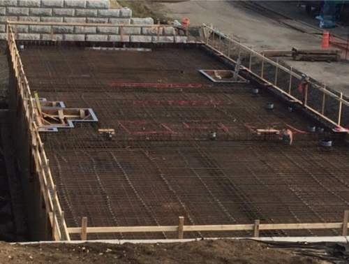 Chantier de construction avec barres d'armature posées, coffrages en bois et un ouvrier en arrière-plan.
