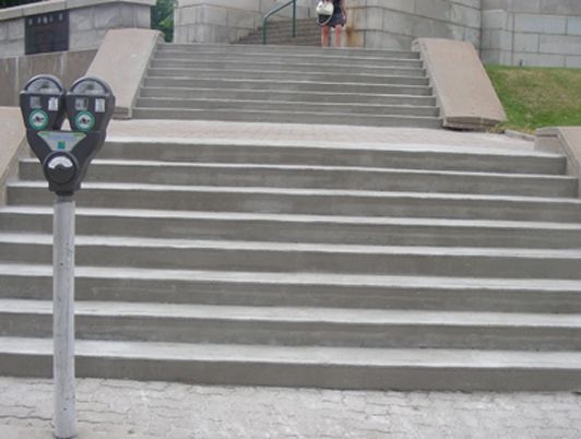 Horodateur devant un large escalier en béton menant à un bâtiment ; une personne en haut.