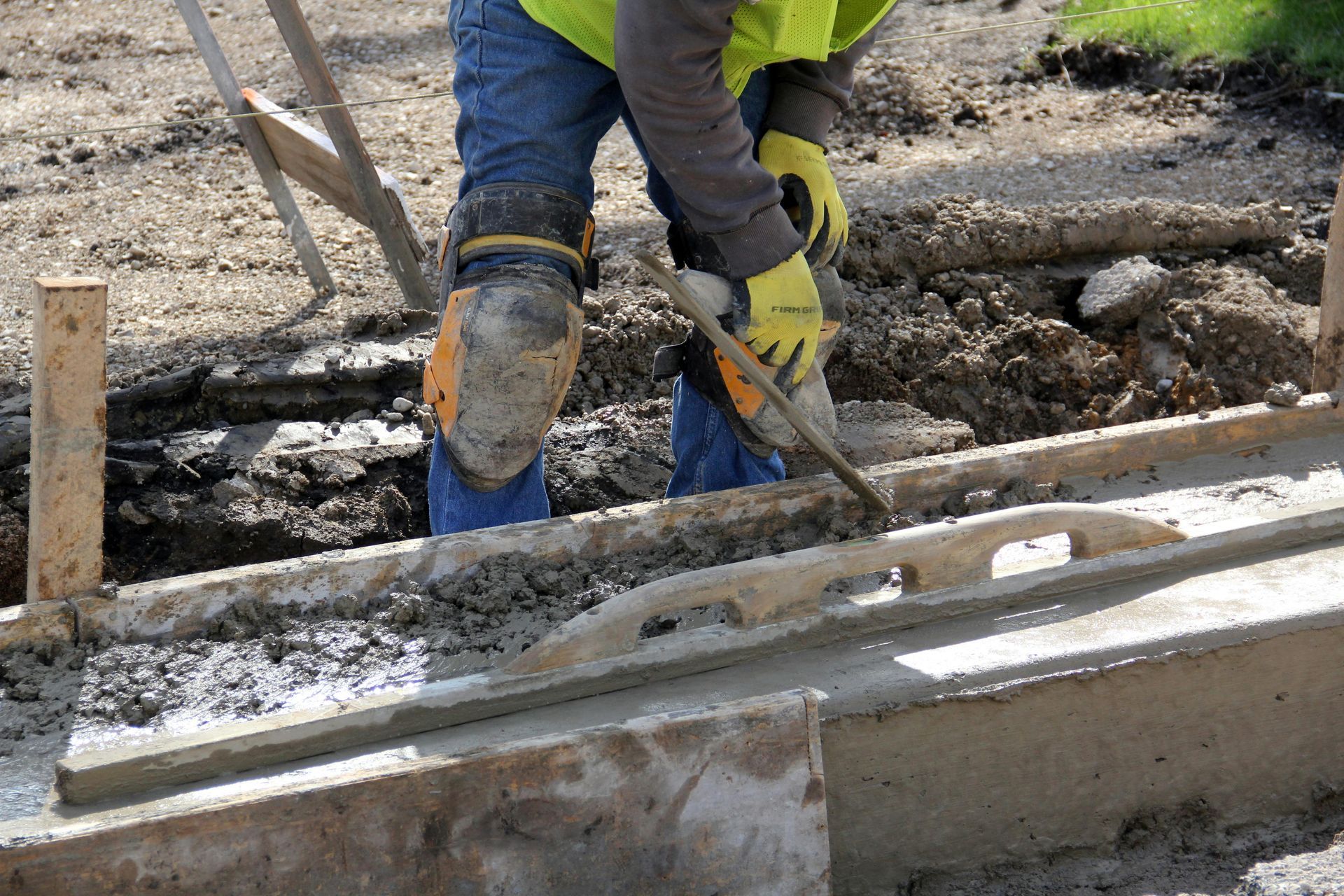 Une personne portant un gilet de sécurité et des gants lisse du béton à la truelle sur un chantier de construction.