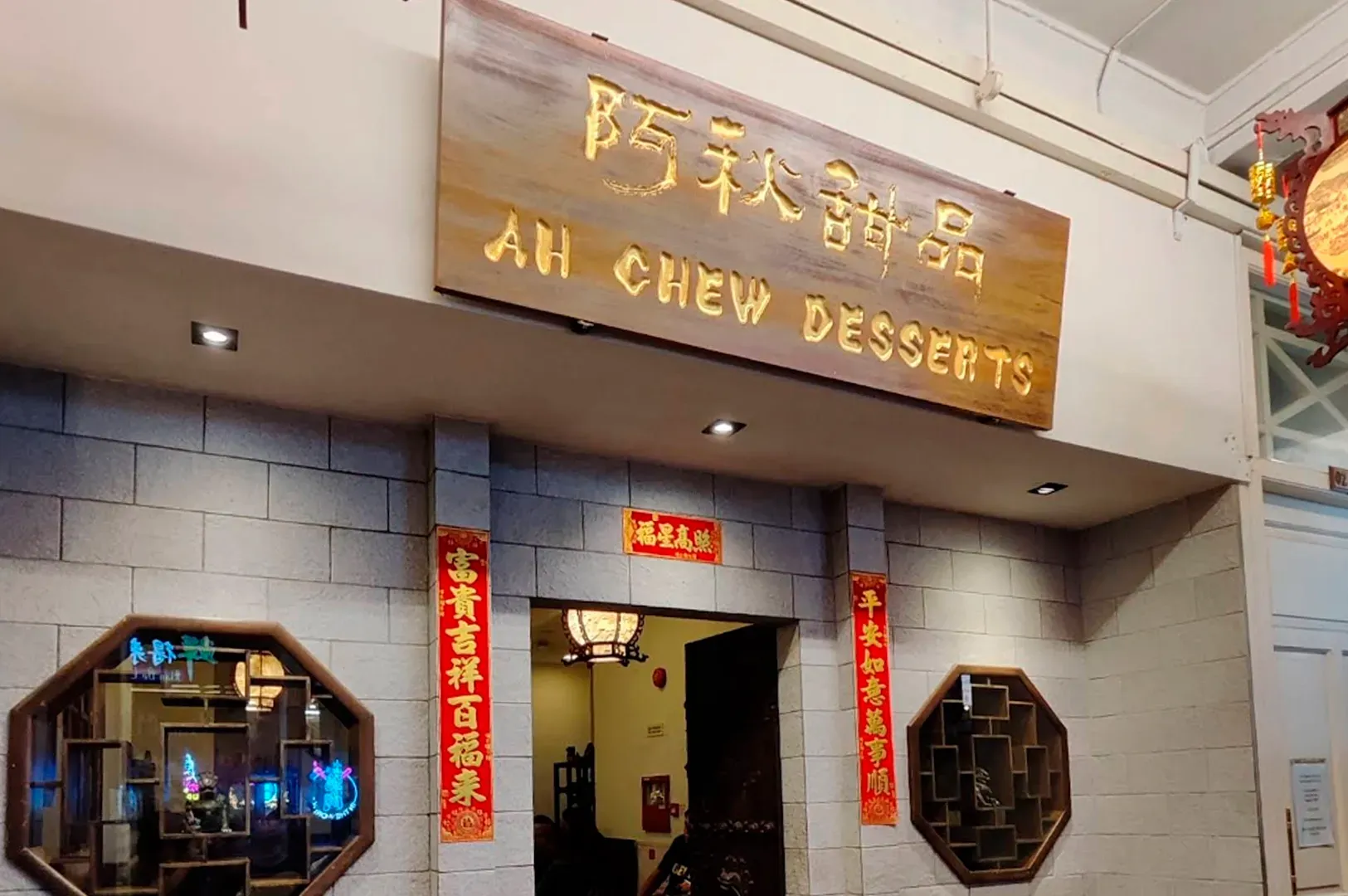 The storefront of Ah Chew Desserts featuring a prominent, rustic wooden signboard with the shop's name carved in gold Chinese calligraphy and English letters. Below the sign, the grey stone wall is decorated with red lunar New Year scrolls. Two octagonal windows with intricate wooden frames flank the entrance, revealing glimpses of a traditional interior.
