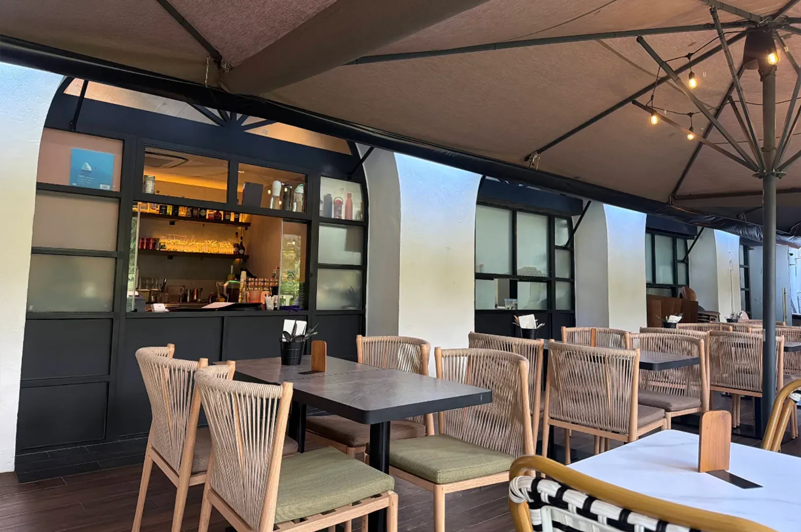 Outdoor restaurant patio with wicker chairs, tables, and a bar under large beige umbrellas