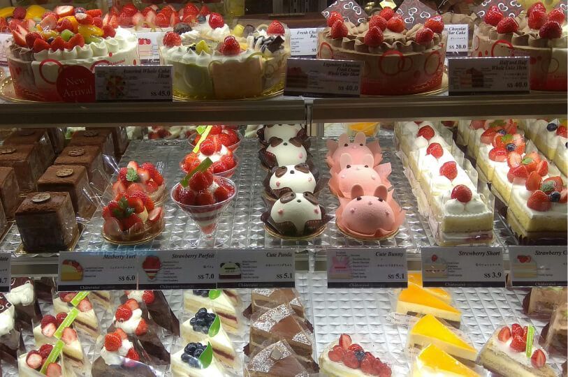 A pastry display case filled with various Japanese-style cakes and desserts, featuring strawberry shortcakes, chocolate cakes, fruit parfaits, and character-themed treats like