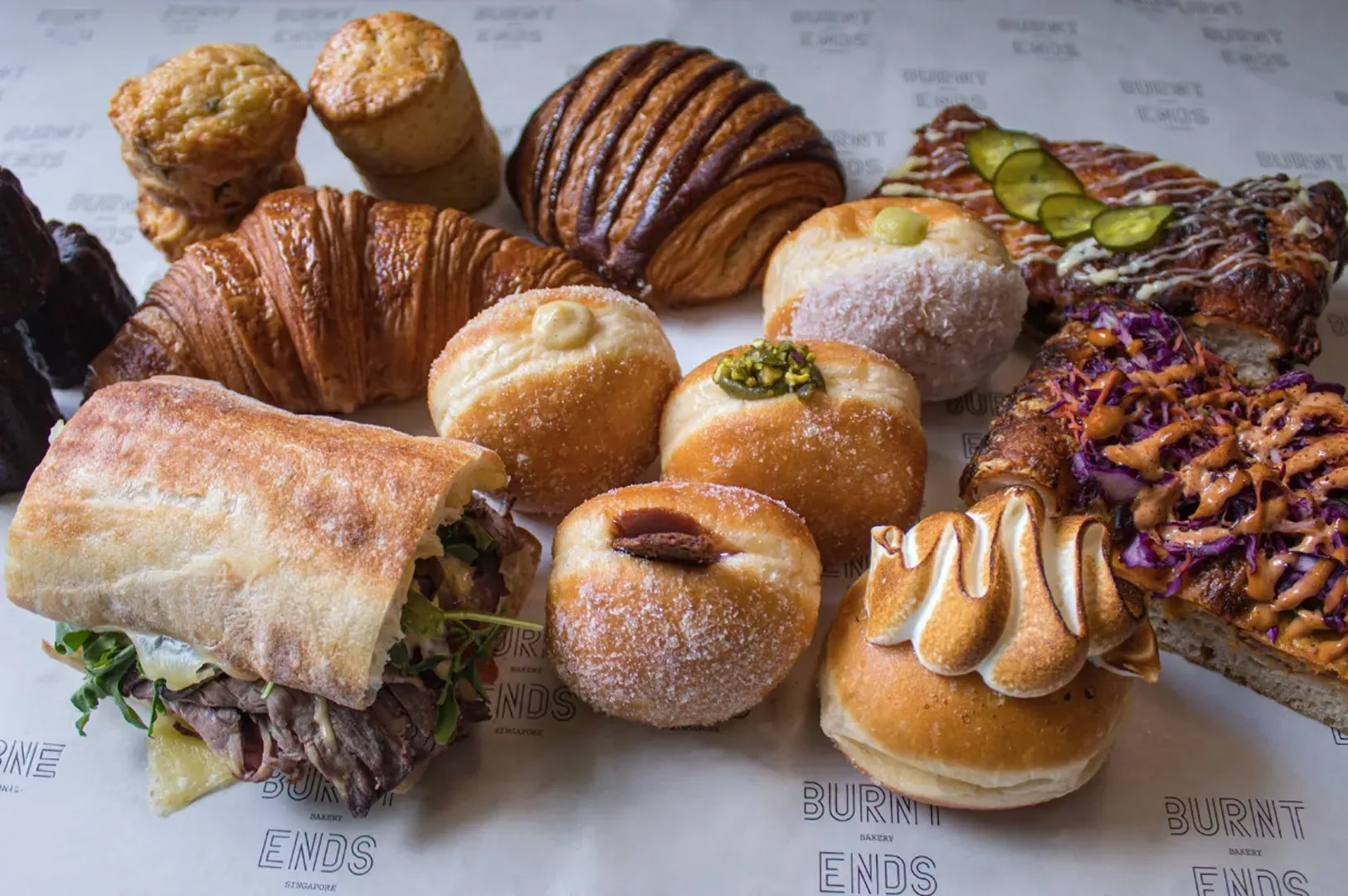 Assorted pastries and bread on a paper-lined table, including croissants, rolls, buns, and a sliced loaf.