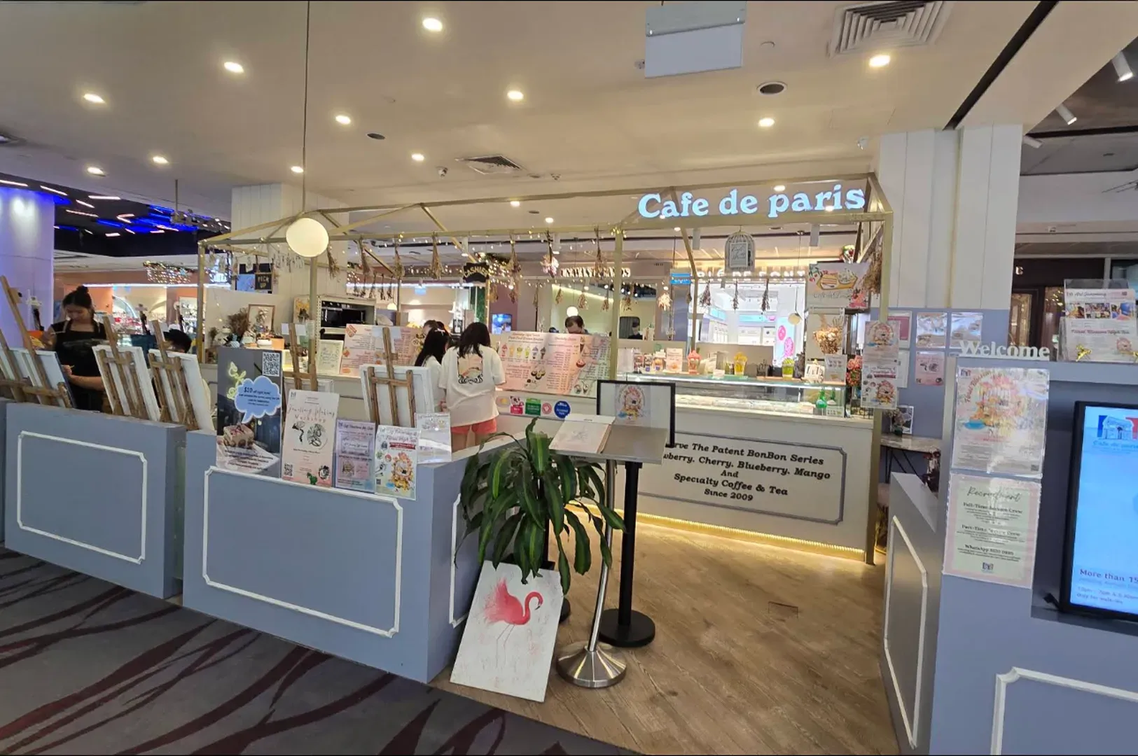 Cafe de Paris bakery kiosk in a mall, with pastries, displays, and a lit sign.