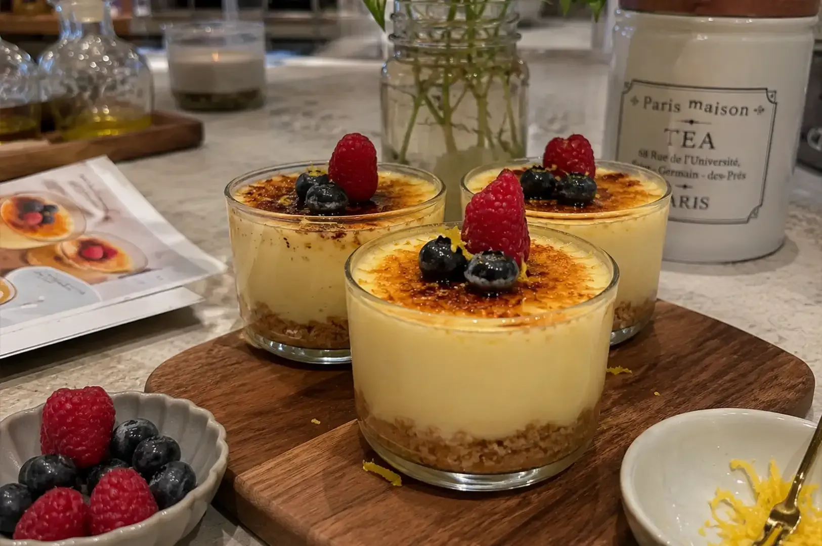 Three berry-topped custards on a wooden board with berries and tea on a table