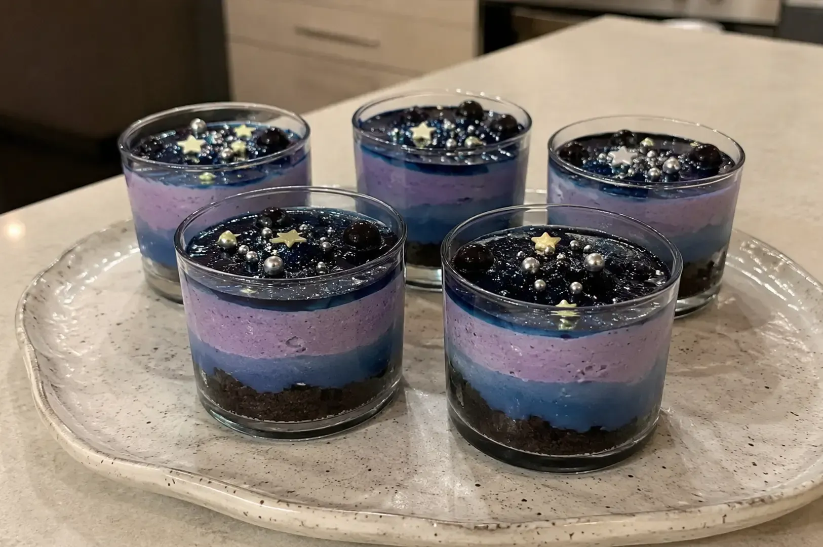 Five layered purple-blue desserts in clear glasses on a white platter