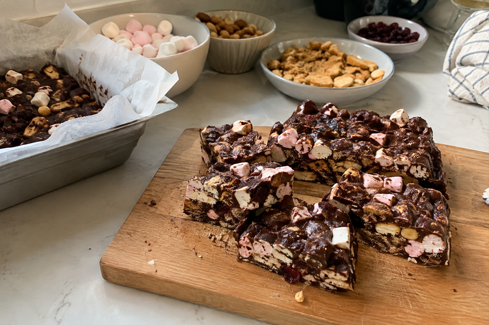 Chocolate bark squares on a cutting board, with bowls of marshmallows, nuts, and dried fruit in the background