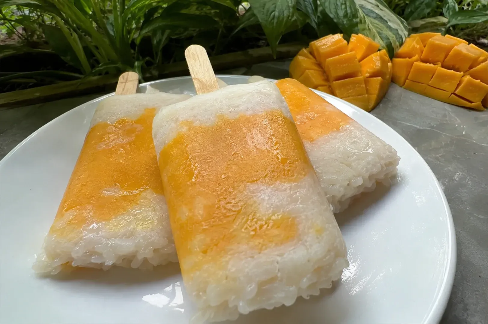 Three coconut rice popsicles on a white plate with sliced mango in the background