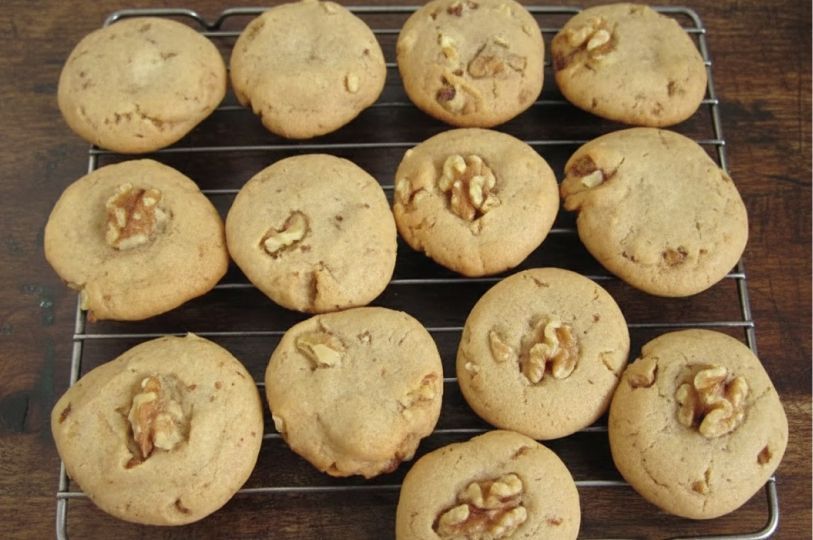 Twelve round, golden-brown honey walnut cookies cooling on a wire rack. Each cookie is topped with a large walnut piece, showing a soft, chewy texture and slightly cracked surface.