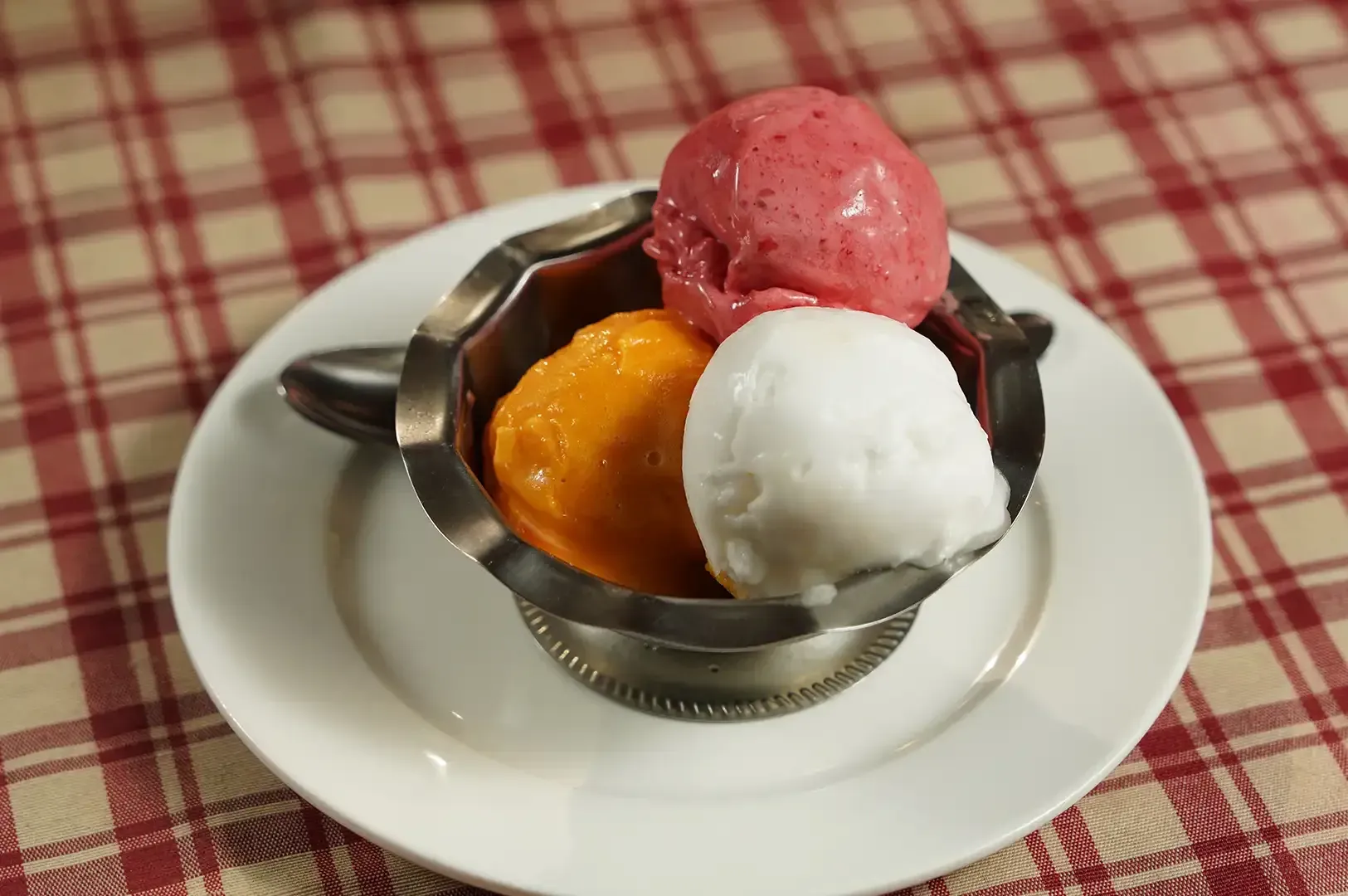 Three scoops of sorbet in a metal bowl on a white plate, with a checkered tablecloth background