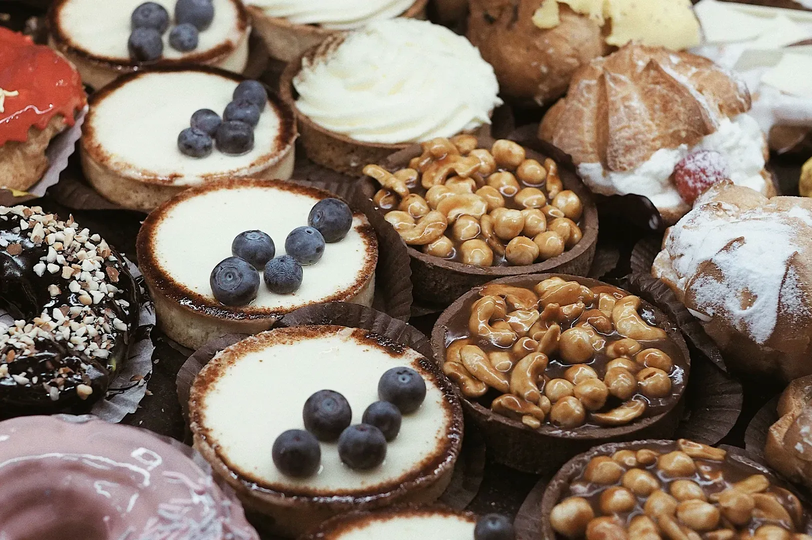 A high-angle close-up of an assortment of gourmet pastries arranged closely together. Visible treats include several small fruit tarts topped with fresh blueberries, rich chocolate tarts generously covered in whole roasted cashews, and a larger pastry topped with white cream and a strawberry.