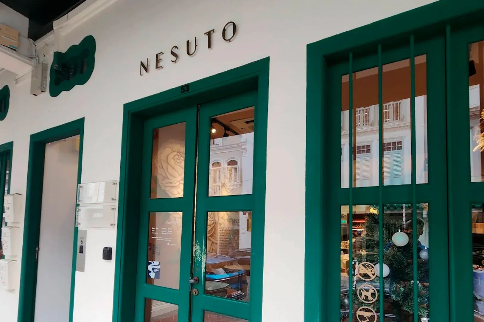 A street-level view of the Nesuto storefront, featuring a clean white facade with vibrant teal-painted door and window frames. The store name is mounted in elegant, thin gold lettering above the main entrance. Large glass panels on the doors and windows reflect the traditional shophouse architecture of the street, while a hint of a warm, decorated interior with a Christmas tree is visible through the right window.