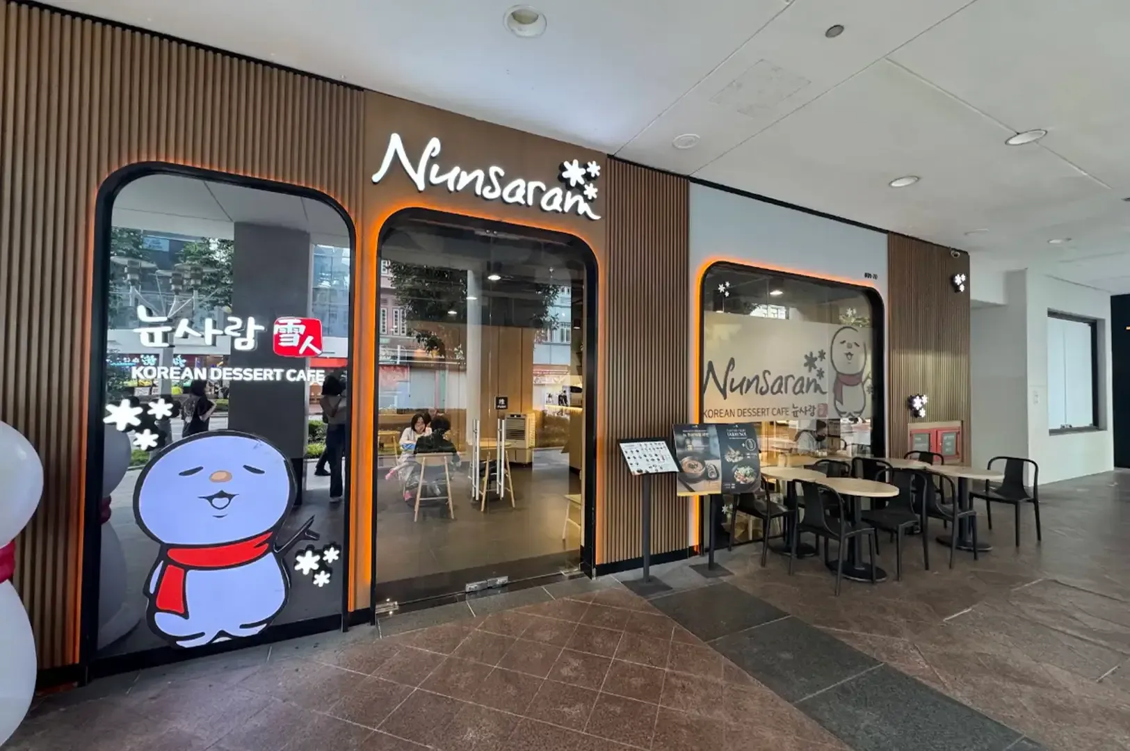Street-side view of a café entrance with glass doors, outdoor tables, and a cartoon character sign.