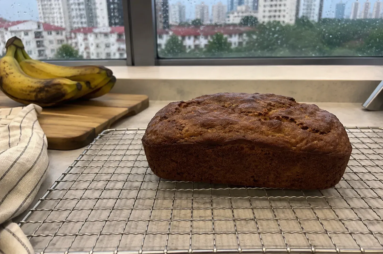 Loaf of banana bread cooling on a rack beside bananas on a kitchen counter by a window